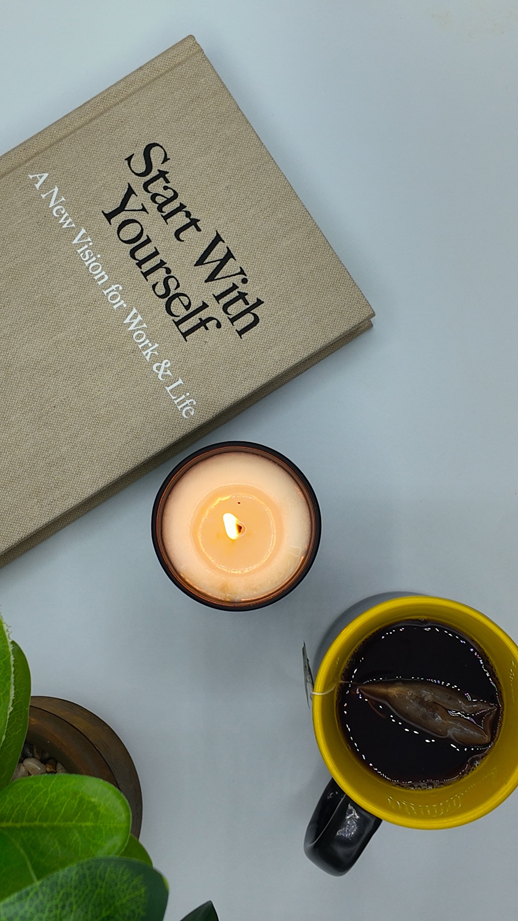 A styled flat-lay featuring a beige hardcover book with the title 'Start With Yourself' on the cover, a lit candle in a dark glass holder, and a yellow mug filled with a dark beverage and a gummy bear, arranged on a light blue-gray background with green leaves in the corner.sleAura Spa & Wellness offers professional massage therapy, facials, pedicures, waxing, nails, brows, lashes, and beauty treatments in Valsayn and Marabella, creating a relaxing luxury spa experience in Trinidad.