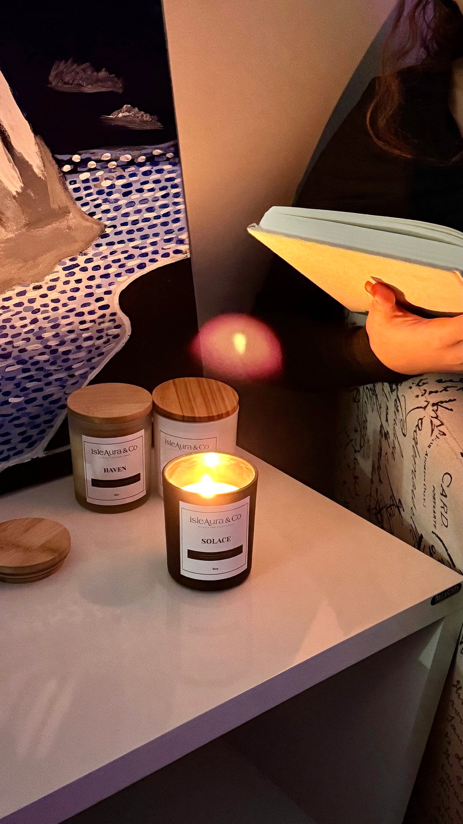 Three soy candles in glass jars with wooden lids are arranged on a white table, with one candle lit and casting a warm glow. A person holding a notebook is partially visible in the background in a dimly lit room.Relaxing lifestyle featuring IsleAura candles, body oils, spa experiences, and wellness rituals designed to promote calm, mindfulness, and intentional self-care in Trinidad